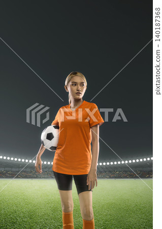 A young Indonesian southeast asian woman soccer or football player in an orange jersey stands on the field of a large stadium at night holding a soccer ball, ready for the game A young Indonesian southeast asian woman soccer or football player in an orange jersey stands on the field of a large stadium at night holding a soccer ball, ready for the game 140187368