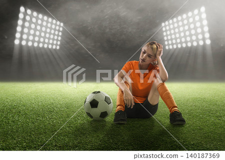An exhausted Indonesian southeast asian female football or soccer player in an orange uniform sits on the turf under bright stadium floodlights, looking dejected and disappointed after a game An exhausted Indonesian southeast asian female football or soccer player in an orange uniform sits on the turf under bright stadium floodlights, looking dejected and disappointed after a game 140187369