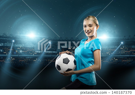 A smiling young Indonesian southeast asian woman in a blue t-shirt stands proudly holding a soccer ball inside a brightly illuminated sports stadium at night, ready for a match A smiling young Indonesian southeast asian woman in a blue t-shirt stands proudly holding a soccer ball inside a brightly illuminated sports stadium at night, ready for a match 140187370
