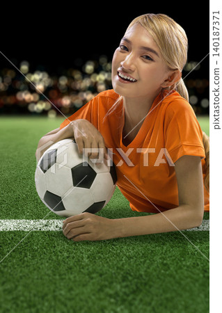 A happy young Indonesian southeast asian woman soccer player in an orange jersey lies on the green pitch, smiling and holding a football at a night stadium A happy young Indonesian southeast asian woman soccer player in an orange jersey lies on the green pitch, smiling and holding a football at a night stadium 140187371