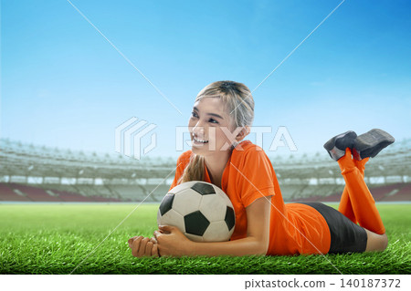 A cheerful young Indonesian southeast asian woman in an orange jersey lies on stadium grass holding a soccer ball A cheerful young Indonesian southeast asian woman in an orange jersey lies on stadium grass holding a soccer ball 140187372