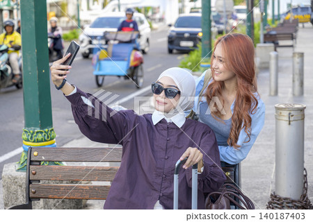 A young Indonesian southeast asian muslim woman in a hijab and her friend smile while taking a selfie with a smartphone on a city sidewalk during their vacation. Travel or vacation concept A young Indonesian southeast asian muslim woman in a hijab and her friend smile while taking a selfie with a smartphone on a city sidewalk during their vacation. Travel or vacation concept 140187403