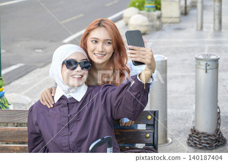 A young Indonesian southeast asian muslim woman in a hijab and her friend smile while taking a selfie with a smartphone on a city sidewalk during their vacation. Travel or vacation concept 140187404