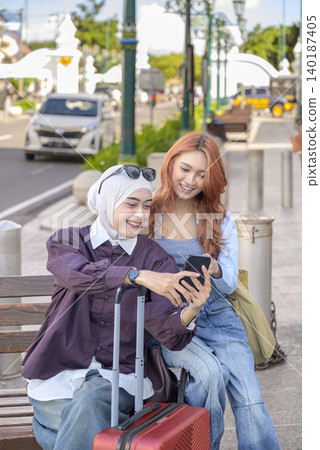 Two young Indonesian southeast asian women, one wearing a hijab, sit on a street bench with a suitcase, smiling while looking at a smartphone together during their travels. Travel or vacation concept Two young Indonesian southeast asian women, one wearing a hijab, sit on a street bench with a suitcase, smiling while looking at a smartphone together during their travels. Travel or vacation concept 140187405