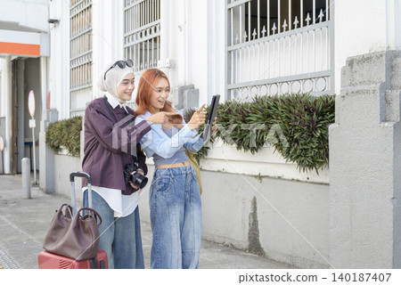 Two young Indonesian southeast asian diverse women, one wearing a hijab, use a digital tablet to find their way on an urban street while on vacation. Travel or vacation concept Two young Indonesian southeast asian diverse women, one wearing a hijab, use a digital tablet to find their way on an urban street while on vacation. Travel or vacation concept 140187407