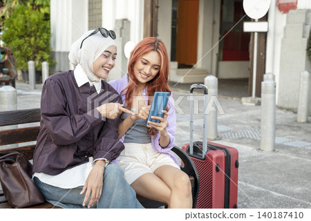 Two young Indonesian southeast asian women, one wearing a hijab, sitting on a bench next to a suitcase and smiling while looking at a smartphone screen together. Travel or vacation concept Two young Indonesian southeast asian women, one wearing a hijab, sitting on a bench next to a suitcase and smiling while looking at a smartphone screen together. Travel or vacation concept 140187410