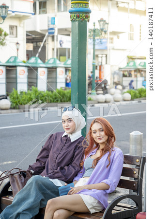 Two stylish young Indonesian southeast asian women, one wearing a hijab and the other with dyed hair, sit on a wooden bench, smiling at the camera. Travel or vacation concept Two stylish young Indonesian southeast asian women, one wearing a hijab and the other with dyed hair, sit on a wooden bench, smiling at the camera. Travel or vacation concept 140187411