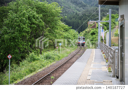 一列單節車廂的本地鐵路列車駛入綠樹環繞的本地車站月台。 一列單節車廂的本地鐵路列車駛入綠樹環繞的本地車站月台。 140187727