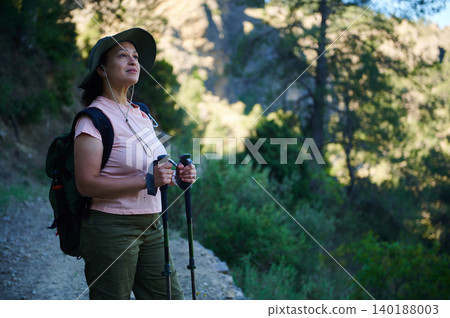 Woman hiking with trekking poles and backpack enjoying nature on a forest trail at sunrise 140188003