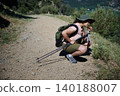 Woman hiking on a sunny mountain trail pausing to smell plants with backpack and trekking poles 140188007