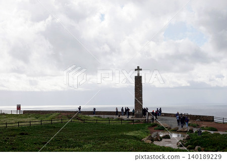 Stone monument with a cross on a coastal cliff overlooking the ocean with tourists under a cloudy sky Stone monument with a cross on a coastal cliff overlooking the ocean with tourists under a cloudy sky 140189229