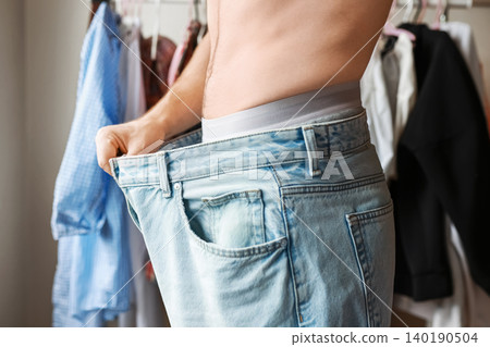 a man shows his weight loss and body transformation by wearing oversized jeans. A concept of healthy lifestyle, diet, and fitness success a man shows his weight loss and body transformation by wearing oversized jeans. A concept of healthy lifestyle, diet, and fitness success 140190504