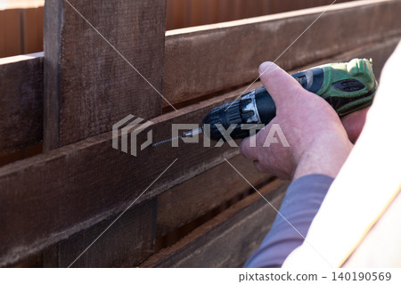man's hands using a green and black cordless electric drill to screw a self-tapping screw into a wooden fence board. The scene captures the practicality of DIY home improvement, woodworking, or constr man's hands using a green and black cordless electric drill to screw a self-tapping screw into a wooden fence board. The scene captures the practicality of DIY home improvement, woodworking, or constr 140190569
