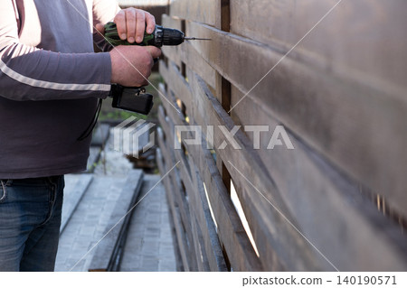 man's hands using a green and black cordless electric drill to screw a self-tapping screw into a wooden fence board. The scene captures the practicality of DIY home improvement, woodworking, or constr man's hands using a green and black cordless electric drill to screw a self-tapping screw into a wooden fence board. The scene captures the practicality of DIY home improvement, woodworking, or constr 140190571