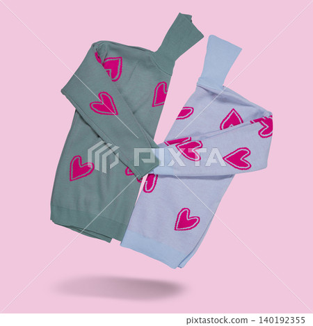 Two cozy knitted turtleneck sweaters with pink heart patterns in grey and light blue colors flying on pastel pink background, copy space. Two cozy knitted turtleneck sweaters with pink heart patterns in grey and light blue colors flying on pastel pink background, copy space. 140192355