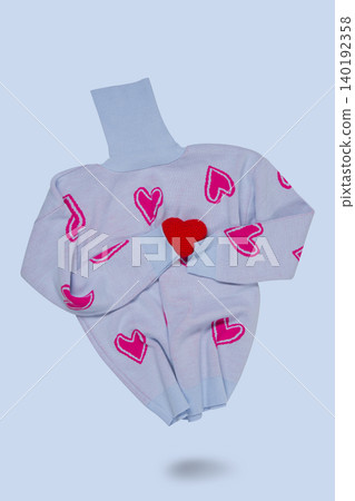 Light blue knitted turtleneck sweater with pink hearts print hugging a small red handmade heart, levitating on blue background. Light blue knitted turtleneck sweater with pink hearts print hugging a small red handmade heart, levitating on blue background. 140192358