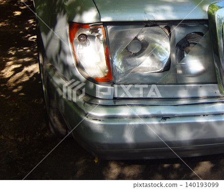 Close-up of a damaged silver car broken front headlight, smashed glass, a cracked indicator lens, and a dented bumper after a road accident 140193099