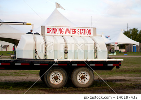 A white mobile drinking water station tank trailer parked at an outdoor festival field with white event tents A white mobile drinking water station tank trailer parked at an outdoor festival field with white event tents 140197432