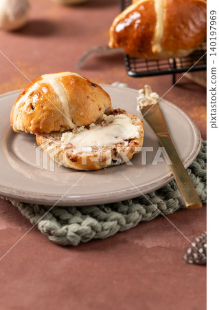Vertical shot of a sliced hot cross bun with a thick layer of butter on a grey plate with a gold knife on a rustic table with spring decorations Vertical shot of a sliced hot cross bun with a thick layer of butter on a grey plate with a gold knife on a rustic table with spring decorations 140197969