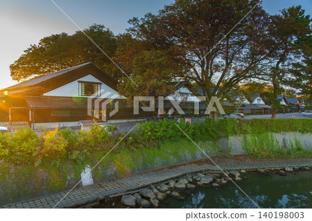 Sakata, Yamagata, Japan - Oct 5 2024, Panoramic view from water of Sankyo Soko Storehouse with an alley of trees nearby, at sunset, without people, Sakata, Japan 140198003