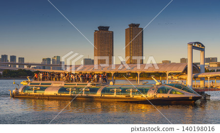 Tokyo, Japan - Oct 13 2024, Panoramic view of Hotaluna water bus filled with passengers sailing along bay, on background of a sharp odaiba with two identical skyscrapers, in evening, Tokyo, Japan Tokyo, Japan - Oct 13 2024, Panoramic view of Hotaluna water bus filled with passengers sailing along bay, on background of a sharp odaiba with two identical skyscrapers, in evening, Tokyo, Japan 140198016
