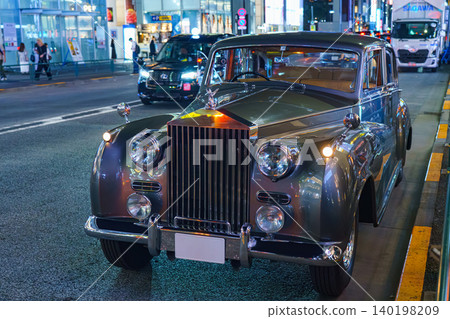 Tokyo, Japan - Oct 15 2024, A close-up view of a vintage Rolls-Royce Silver Cloud car parked at curb in central Tokyo, with people walking in background and cars speeding along road, Tokyo, Japan Tokyo, Japan - Oct 15 2024, A close-up view of a vintage Rolls-Royce Silver Cloud car parked at curb in central Tokyo, with people walking in background and cars speeding along road, Tokyo, Japan 140198209