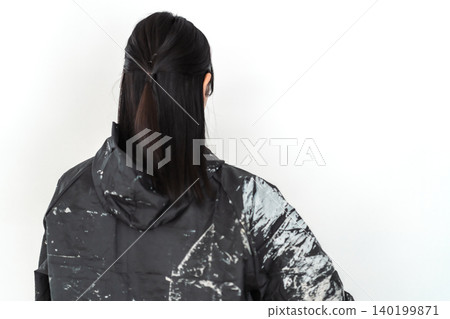 A smiling young female worker wearing paint-stained work clothes and holding a helmet against a white background. 140199871