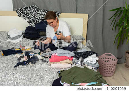 A 52-year-old woman sits on her bed, sorting a pile of laundry. Clothes are spread out around her as she carefully organizes socks and other garments in her bedroom A 52-year-old woman sits on her bed, sorting a pile of laundry. Clothes are spread out around her as she carefully organizes socks and other garments in her bedroom 140201939