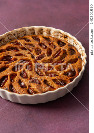 A plum pie is placed on a table with thinly sliced plums visible on top. Bowls of fresh plums are nearby with empty plates waiting for dessert 140201950