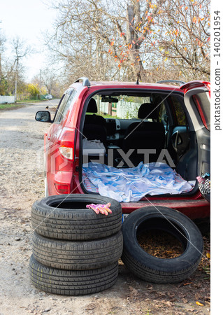 A red car sits along a gravel road as summer tires are removed and replaced with winter tires. Tools and a blanket are on the back seat. It is fall, and leaves are changing color 140201954