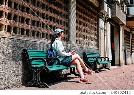 Mature 50 years old Russian woman tourist in cap sitting on green bench on street holding phone during solo travel summer vacation in Macau China Mature 50 years old Russian woman tourist in cap sitting on green bench on street holding phone during solo travel summer vacation in Macau China 140202413