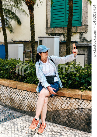 Mature 50 years old Russian woman tourist in cap taking selfie with phone near Sir Robert Ho Tung Library building during summer vacation in Macau Mature 50 years old Russian woman tourist in cap taking selfie with phone near Sir Robert Ho Tung Library building during summer vacation in Macau 140202414