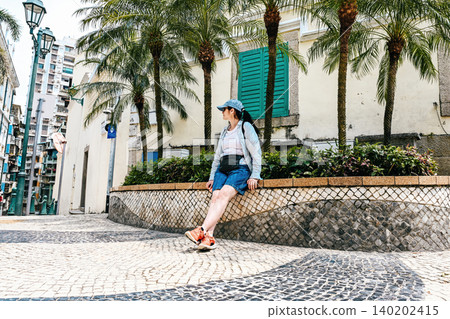 Mature 50 years old Russian woman tourist in cap relaxing under palm trees near Sir Robert Ho Tung Library during summer vacation in Macau China Mature 50 years old Russian woman tourist in cap relaxing under palm trees near Sir Robert Ho Tung Library during summer vacation in Macau China 140202415