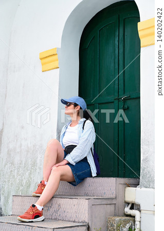 Mature 50 years old Russian woman tourist in cap sitting on stairs near ancient green door of Guia Lighthouse during summer vacation in Macau China 140202422