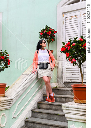 Mature 50 years old Russian woman tourist brunet standing on stairs near mint green house built in 1921 with camellia flowers in Taipa Houses Macau Mature 50 years old Russian woman tourist brunet standing on stairs near mint green house built in 1921 with camellia flowers in Taipa Houses Macau 140202426