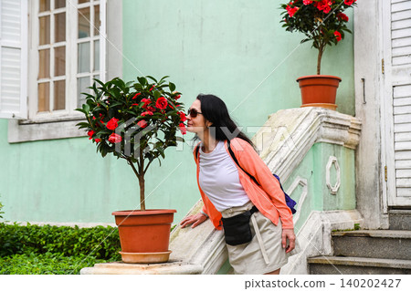 Mature 50 years old Russian woman tourist brunette smelling red camellia flower on stairs near mint green house built in 1921 at Taipa Houses Macau Mature 50 years old Russian woman tourist brunette smelling red camellia flower on stairs near mint green house built in 1921 at Taipa Houses Macau 140202427
