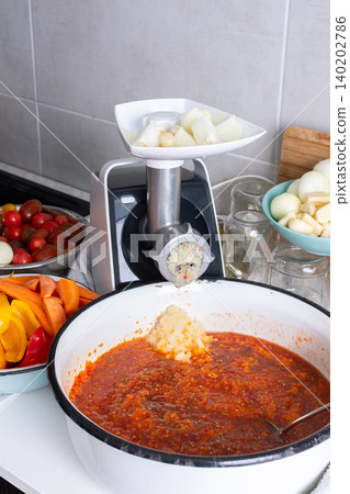 Fresh vegetables are being ground to prepare Adjika in a kitchen. Bright carrots, tomatoes, and peppers are added to a large bowl for a spicy Georgian seasoning Fresh vegetables are being ground to prepare Adjika in a kitchen. Bright carrots, tomatoes, and peppers are added to a large bowl for a spicy Georgian seasoning 140202786