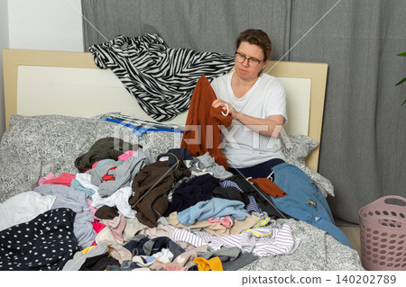 A woman, age 52, sits on her bed in a bedroom while sorting through clean laundry. She holds a piece of clothing in her hands as she organizes her belongings on the bed A woman, age 52, sits on her bed in a bedroom while sorting through clean laundry. She holds a piece of clothing in her hands as she organizes her belongings on the bed 140202789