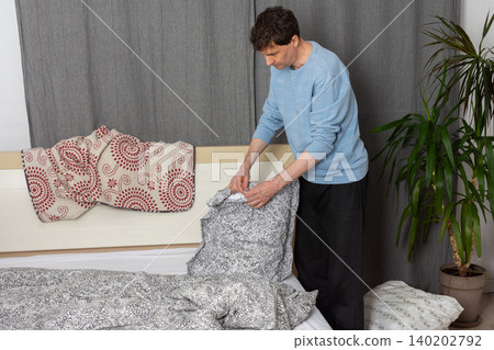 A 53-year-old man is changing the bed linen in his bedroom. He holds a pillow while arranging the sheets and blankets, focusing on the task of keeping his space tidy A 53-year-old man is changing the bed linen in his bedroom. He holds a pillow while arranging the sheets and blankets, focusing on the task of keeping his space tidy 140202792