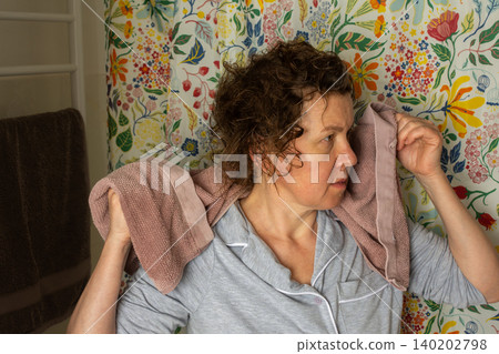 A 50-year-old woman prepares for her day in her bathroom. She is focused while removing a towel from her hair. The room is bright and colorful, reflecting a cheerful atmosphere A 50-year-old woman prepares for her day in her bathroom. She is focused while removing a towel from her hair. The room is bright and colorful, reflecting a cheerful atmosphere 140202798