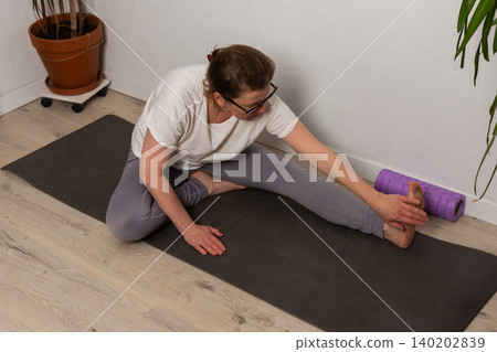 A 52-year-old woman is exercising at home on a yoga mat. She stretches her legs and uses a foam roller. The room is simple and has basic decor, making it a workout space A 52-year-old woman is exercising at home on a yoga mat. She stretches her legs and uses a foam roller. The room is simple and has basic decor, making it a workout space 140202839