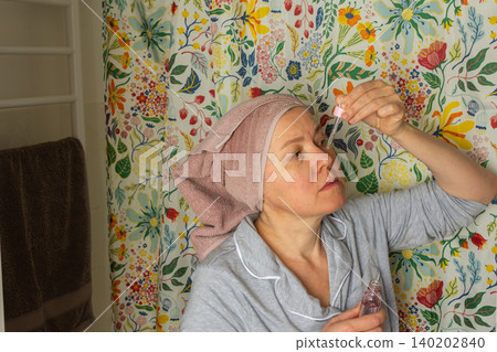 A woman in her fifties is in the bathroom applying facial serum. She wears a towel on her head and soft pajamas. It is morning, and colorful patterns surround her 140202840