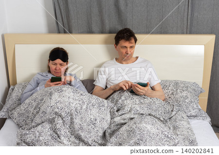 A fifty-year-old couple wakes up in their bedroom. Both are in bed with blankets. They are looking at their phones in the quiet of the morning, enjoying a relaxed start to the day A fifty-year-old couple wakes up in their bedroom. Both are in bed with blankets. They are looking at their phones in the quiet of the morning, enjoying a relaxed start to the day 140202841