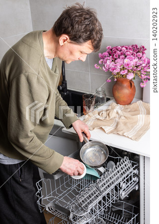 A man is putting dirty dishes into the dishwasher in his kitchen. He focuses on the task while several items rest in the appliance, ready for cleaning 140202923