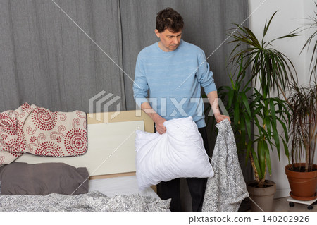 A 53-year-old man replaces the bed linen in his bedroom. He gathers clean sheets and works on making the bed comfortable for sleeping A 53-year-old man replaces the bed linen in his bedroom. He gathers clean sheets and works on making the bed comfortable for sleeping 140202926