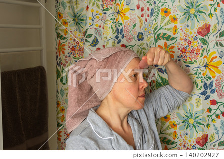 A 50-year-old woman follows her morning routine in the bathroom. She applies skincare products on her face while wearing a towel on her head and comfortable clothing A 50-year-old woman follows her morning routine in the bathroom. She applies skincare products on her face while wearing a towel on her head and comfortable clothing 140202927