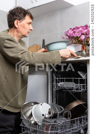 A man stands in his kitchen taking clean dishes out of the dishwasher. He focuses on the process of unloading the clean dishes and organizing them for use A man stands in his kitchen taking clean dishes out of the dishwasher. He focuses on the process of unloading the clean dishes and organizing them for use 140202928