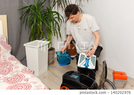 A 53-year-old man is getting ready to clean by preparing a vacuum cleaner. He focuses on the vacuum's parts in a plain room with plants nearby A 53-year-old man is getting ready to clean by preparing a vacuum cleaner. He focuses on the vacuum's parts in a plain room with plants nearby 140202976