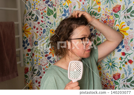 A 50-year-old woman holds a hairbrush while looking in the mirror during her morning bathroom routine. She checks her hair near a floral shower curtain 140203077