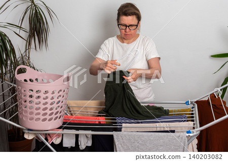 A 50-year-old woman with glasses hangs washed clothes on a drying rack in her indoor space. She sorts through the laundry from a pink basket as she prepares it to dry A 50-year-old woman with glasses hangs washed clothes on a drying rack in her indoor space. She sorts through the laundry from a pink basket as she prepares it to dry 140203082
